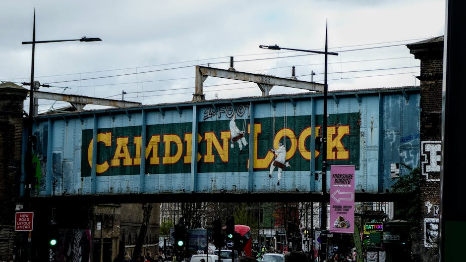 The image depicts a busy urban scene at Camden Market with a prominent overhead sign reading 'Camden Lock' in bold yellow and red letters. The sign is mounted on a metal framework spanning across a st