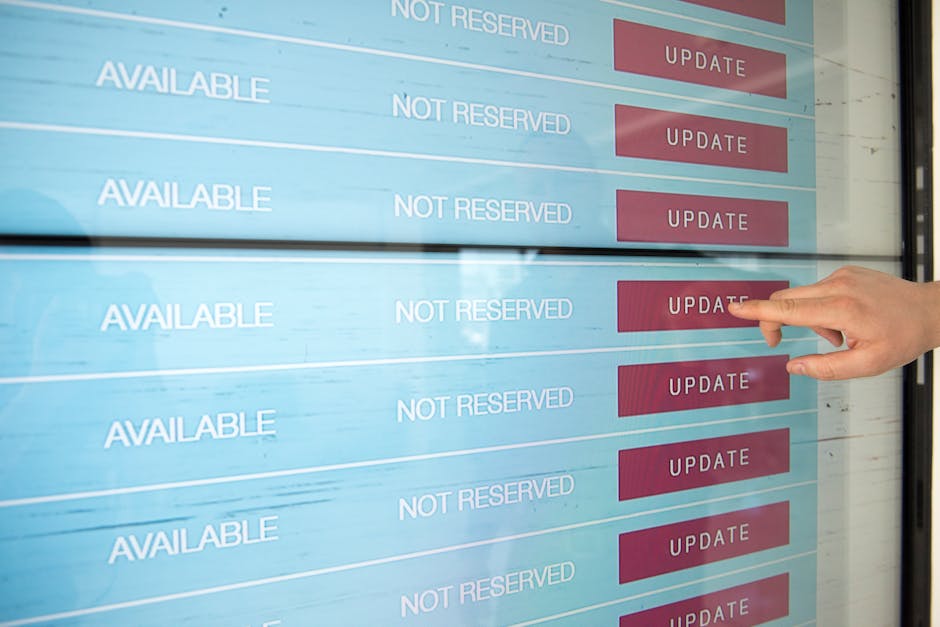 A person's hand pointing to an electronic screen displaying scheduling options with the word 'AVAILABLE' in white text and 'NOT RESERVED' in light blue, alongside red 'UPDATE' buttons. The screen is p
